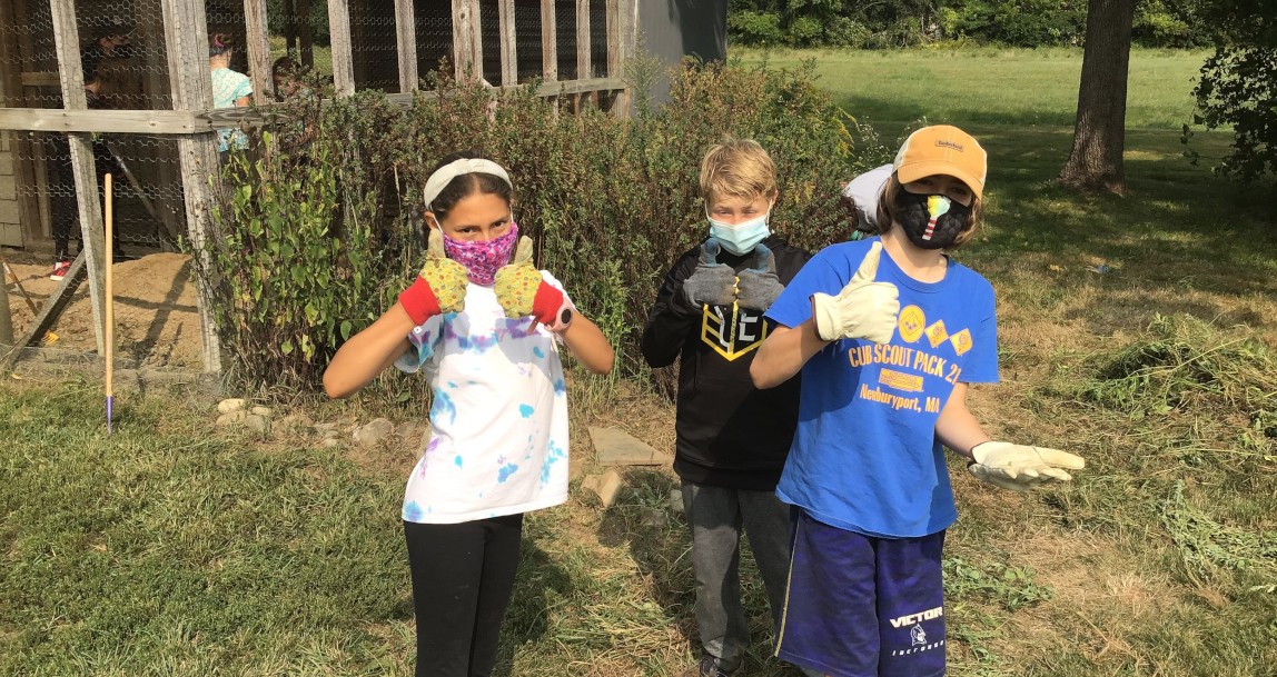 3 students wearing masks give a thumbs up in front of the pollinator garden and chicken coop at SPL