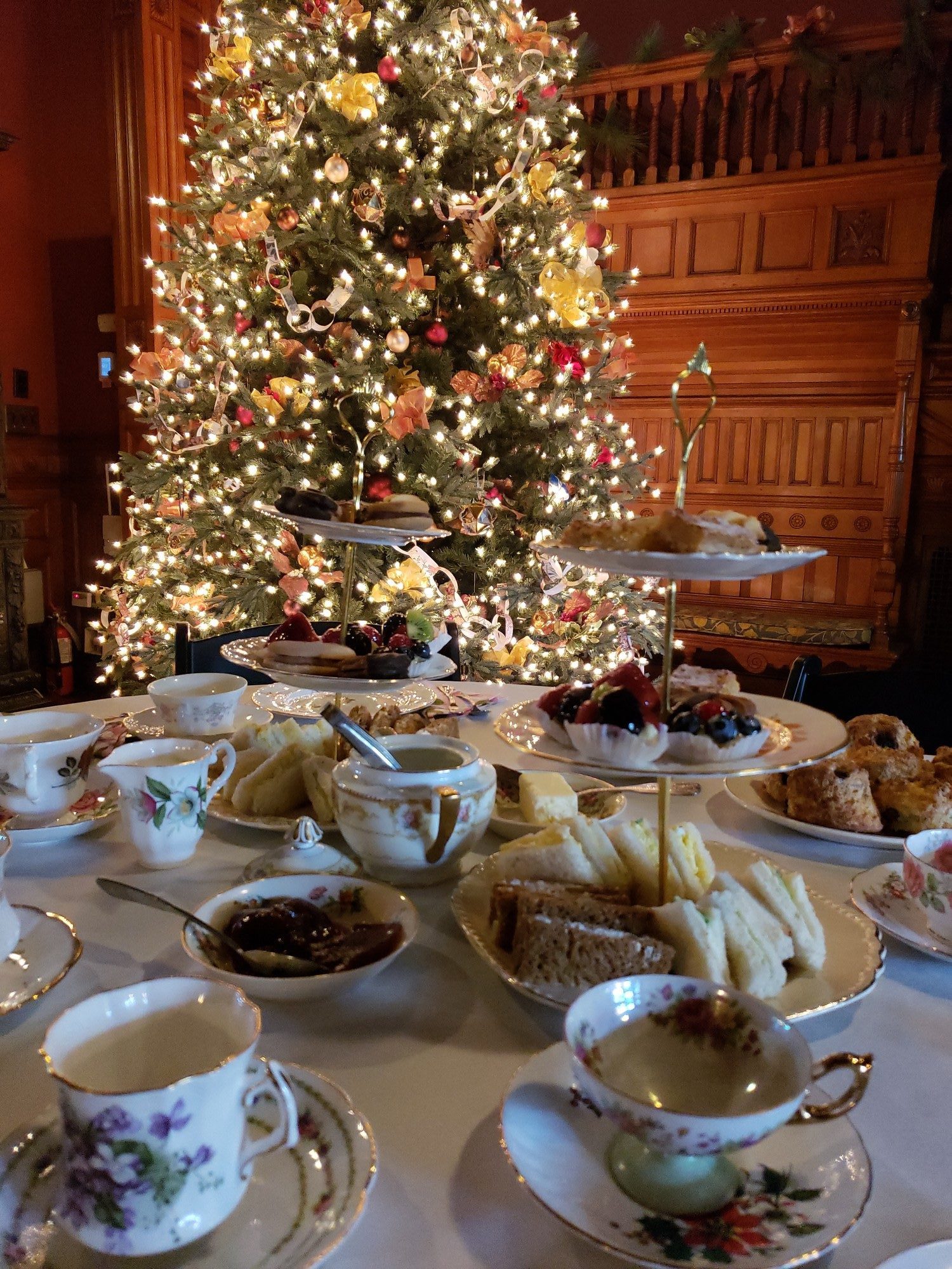 A table with teacups, finger sandwiches, and desserts with a Christmas tree in the background.