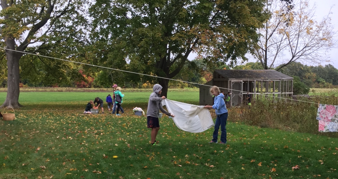 In the foreground, two students prepare to hang a wet sheet on a laundry line. In the background, students work with washboards over buckets, doing the laundry 